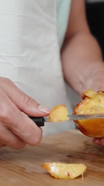 Beyaz önlüklü bir kadın, ahşap bir kesme tahtasıyla nektarini yavaş çekimde kesiyor. Dikey video.