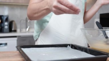 Beyaz önlüklü kadın, mutfağın arka planında, un serpiştirip parşömen kağıdına serpiyor. yavaş çekim.
