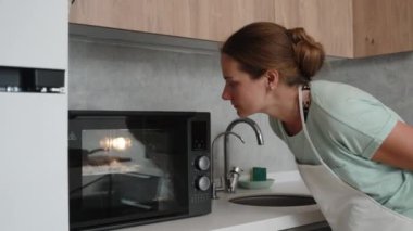 Mutfaktaki beyaz önlüklü genç kadın kremalı kekin nasıl piştiğine bakmak için fırına bakıyor..