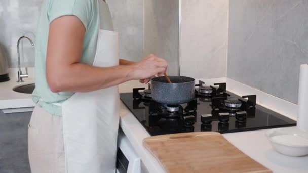 在一间明亮的厨房里 一位女士穿着白色围裙 慢吞吞地在煤气炉上的平底锅里搅拌着什么东西 — 图库视频影像
