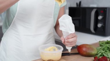 Beyaz önlüklü pasta şefi pasta torbasını kremayla dolduruyor. Masada mango ve nane yaprakları var..
