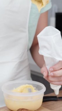 Beyaz önlüklü pasta şefi pasta torbasını kremayla dolduruyor. Masada mango ve nane yaprakları var. Dikey video.