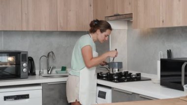 Beyaz önlüklü bir kadın gaz sobasının yanında duruyor çırpıcıyla bir şey karıştırıyor. Mutfağın parlak bir iç mekanı var. yavaş çekim.