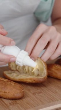Beyaz önlüklü pasta şefi pasta poşetinden eklerine krema sıkıyor. Dikey video.