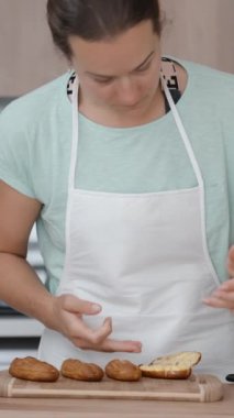 Beyaz önlüklü genç bir kadın pastaları hazırlıyor ve yavaşça parmağını yalıyor. Dikey video.