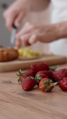 Kadın arka planda yemek pişirirken kamera ön plana odaklanıyor ve ahşap bir masada çilek ve nane yapraklarını gösteriyor. Dikey video.