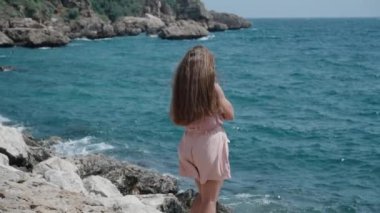 Pembe elbiseli genç bir kadın deniz kenarındaki kayalıklarda duruyor, uzun saçları ve kıyafetleri rüzgarda savruluyor, ağır çekimde..