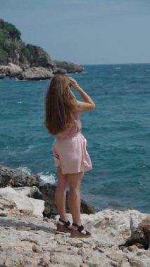 Kısa pembe şortlu genç bir kadın ve uzun saçlarını düzelten rüzgarlı havada deniz kenarındaki kayalıklarda duran bir atlet. Ağır çekim. Dikey video.