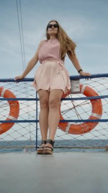 Genç bir kadın geminin üst güvertesinde duruyor, saçları ve şortu rüzgarda dalgalanırken, arka planda can simidi var. Ağır çekim. Dikey video.