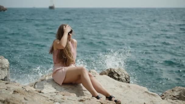 一位穿着粉色短裤的年轻女子坐在岩石上 身后是海浪和水花 她调整她的头发 — 图库视频影像