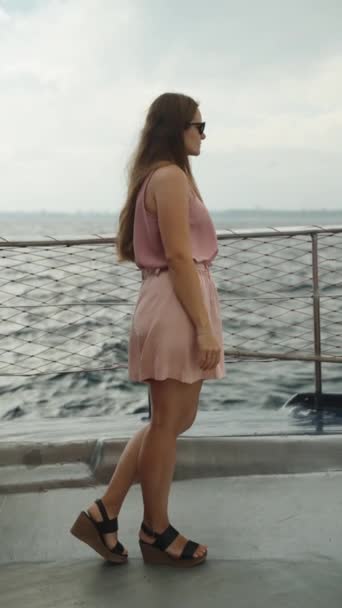 一位身穿粉色短裙和高台凉鞋的年轻女子站在一艘海轮的甲板上 紧紧抓住船的栏杆 在她身后是汹涌的大海 缓慢地移动着 垂直录像 — 图库视频影像