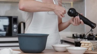 Kadın çırpma aletlerini el blendırına yerleştiriyor ve kaseye beyaz bir şey döküyor..