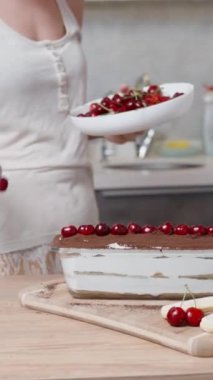 Genç kadın, Tiramisu pastasını bitirmiş bir fotoğraf için masayı kirazlarla süslüyor. Dikey video.