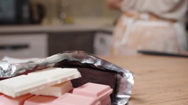 Masanın üzerinde farklı renkte çikolatalar var ve kadın bardağı çikolatayla çıkarıp mikrodalgaya koyuyor..