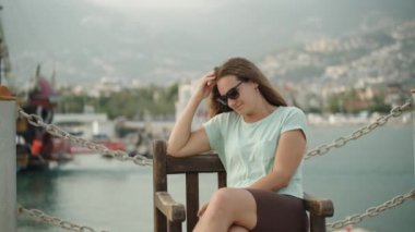 Genç bir kadın deniz kenarında oturur arkasında dağlar ve gemilerle saçlarını düzeltir..