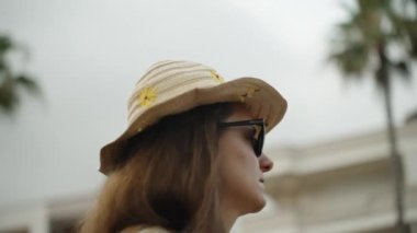 Hasır şapka ve güneş gözlüğü takan genç bir kadın, arka planda palmiye ağaçları ile şehirde geziniyor. Görüntü alçak bir açıdan ve sahne yavaş çekimde..