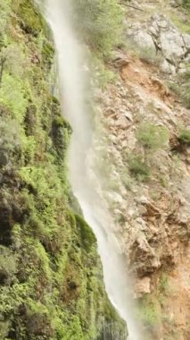 Kayalık arazi ve seyrek bitki örtüsüyle çevrili bir dağ şelalesi. Dikey video.