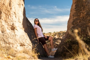 Genç bir kadın kayaların arasında poz veriyor, birine yaslanıyor. Arka planda vadi yarığı var. Güneşli hava.