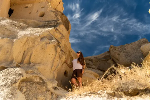 A young woman examines ancient structures in the rocks against the backdrop of wispy clouds in the blue sky.