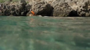 Kamera suyun altına iniyor ve bir şnorkel maskesi takmış genç bir kadını denizin yüzeyinde yüzerken deniz tabanındaki kayalara bakarken yakalıyor..