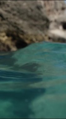 Dikey video. Kamera, su kenarında, turkuaz okyanus dalgalarında, uçurumların arka planında sallanıyor..