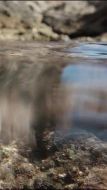 Dikey video. Kayalıklardaki kayalık kıyı şeridi, sudaki kamera iki dünyanın birleşmesini yakalıyor, su ve hava..