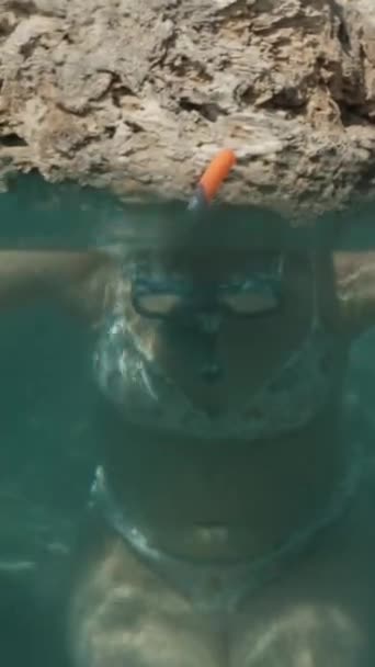 垂直录像 一位年轻女子在海中 在悬崖的簇拥下 调整了潜水面罩 在水下潜水 相机跟着她 — 图库视频影像