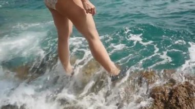 Mayo giymiş genç bir kadın kayalık bir uçurumda özel su ayakkabıları giyerek duruyor ve dalgalar ayaklarına çarpıyor. Yavaş çekim.