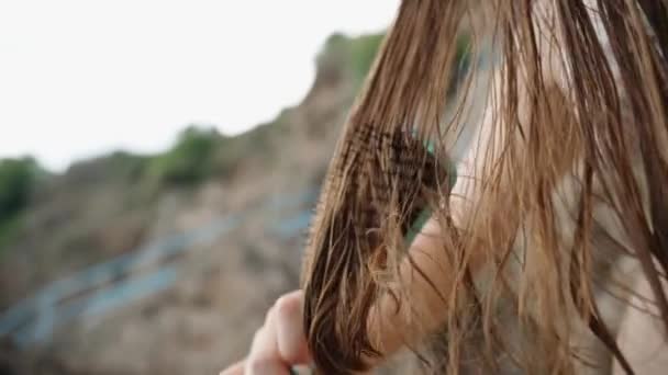 这个女人 有着湿透了的 长长的 浅褐色的游泳头发 慢慢地把她的头发梳理在岩石中靠近大海的地方 镜头是慢镜头中的特写 — 图库视频影像