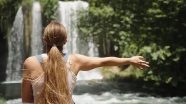 Ağır çekimde, uzun saçlı genç bir kadın ormanda bir dağ nehrindeki şelaleye doğru elini uzatır..