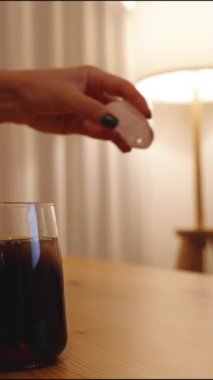 Dikey video. Bir kadın kola bardağına buz küpü atıyor. Ahşap bir masa, arka planda lambadan gelen sıcak bir ışık ve beyaz duvarlar var..