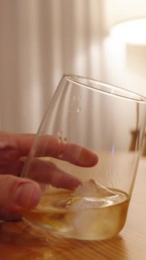 Dikey video. Yalnız bir kadın elindeki viski bardağını çevirir, ahşap bir masanın arkasında oturur, arkasındaki lambadan hafifçe ışık yayılır..