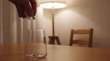 Bir adam, arka plandaki bir lambadan gelen loş ve sıcak ışıkla ahşap bir masanın üzerindeki bir bardağa buz küpü atar..