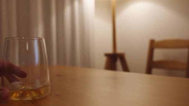 男は木製のテーブルの上に氷でウイスキーのグラスを動かします ランプからの暖かい光がシーンを照らします — ストック動画