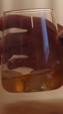 Dikey video. Ağır çekimde, genç bir kadın arka plandaki lambanın ışığıyla viskiyi bardağa karıştırır ve sonra viskiyi burnuna yaklaştırır..