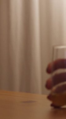 Dikey video. Ahşap bir masada iki bardak viski, ama genç bayan loş lamba ışığında tek başına içiyor..