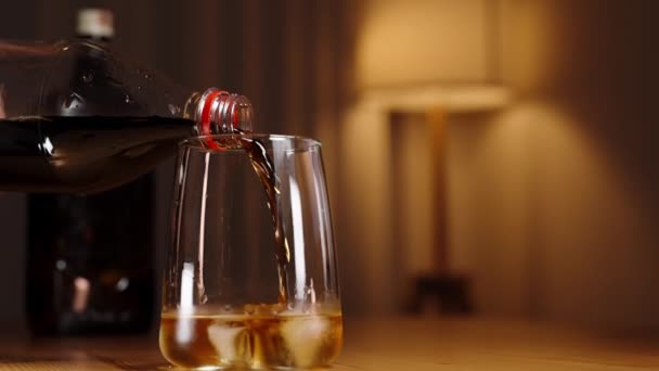 コーラはウイスキーグラスに注ぎ込まれ 背景にボトルが入った木製のテーブルに置かれます 床ランプが部屋に暖かい光を放つ スローモーション — ストック動画