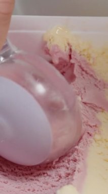 Dikey video. Yuvarlak bir dondurma kepçesi kaptan topluyor, yakın plan, pembe tonlarda..