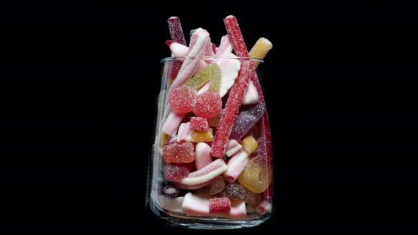 一种透明的玻璃杯 盛满了各种形状和颜色的糖果 上面撒满了糖 它在黑色的背景上旋转 彼此隔离 — 图库视频影像