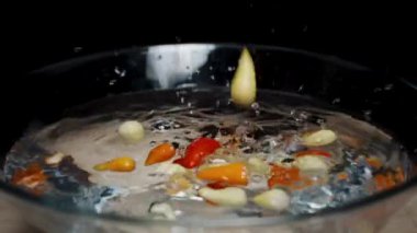 Çok renkli mini kırmızı biberler, siyah arka plana karşı ağır çekimde cam bir kase suya düşüyorlar..