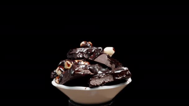 チョコレートとナッツでコーティングされたホワイトボウルの壊れたチョコレートバー 黒い背景に回転 — ストック動画