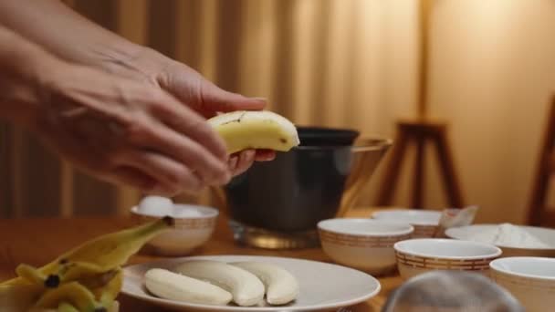 女性がデザートを準備している 彼女はバナナを剥がしている テーブルの上の成分とボウル 背景のランプからの暖かい周囲の光 — ストック動画