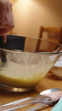Dikey video. Kek hamurunu cam bir kaseye karıştırarak hazırlayan bir kadının yakın plan fotoğrafı. Malzemeler masanın etrafına saçılmış..