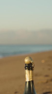 Dikey video. Noel ağacını süslemek için altın parıltılı bir süs topu bir şampanya şişesinin boynunda duruyor. Andolsun gün doğarken deniz kıyısına..