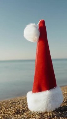 Dikey video. Güneşli bir sabahta deniz kenarında asılı duran kırmızı Noel Baba şapkasının etrafında daireler çiziyor..