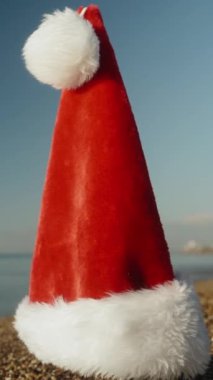 Dikey video. Kırmızı Noel Baba şapkası kumlu sahilde güneşli bir sabahın erken saatlerinde, yakın planda tutulur..