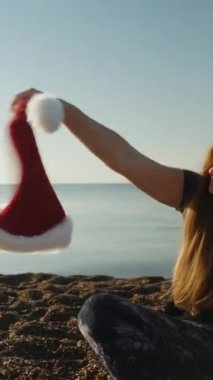 Dikey video. Genç bir kadın sabahın erken saatlerinde sahilde oturur ve elinde kırmızı Noel Baba şapkasını sallar..