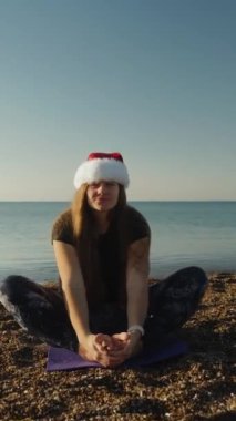 Dikey video. Noel sabahında genç bir kadın kırmızı Noel Baba şapkasıyla denizin kenarında oturur ve şakacı bir şekilde dilini uzatır..