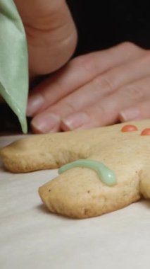 Dikey video. Bir kadın, insan şeklinde zencefilli kurabiye üzerine krema çiziyor. Yakın çekim görünümü.