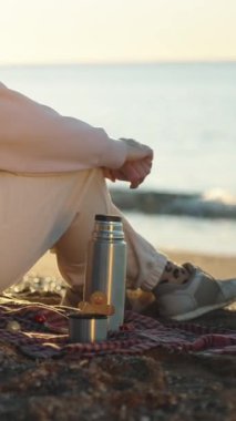 Dikey video. Deniz kenarında serin bir sabahta, bir kadın elinde termos ve battaniyeli zencefilli bir adamla oturur..